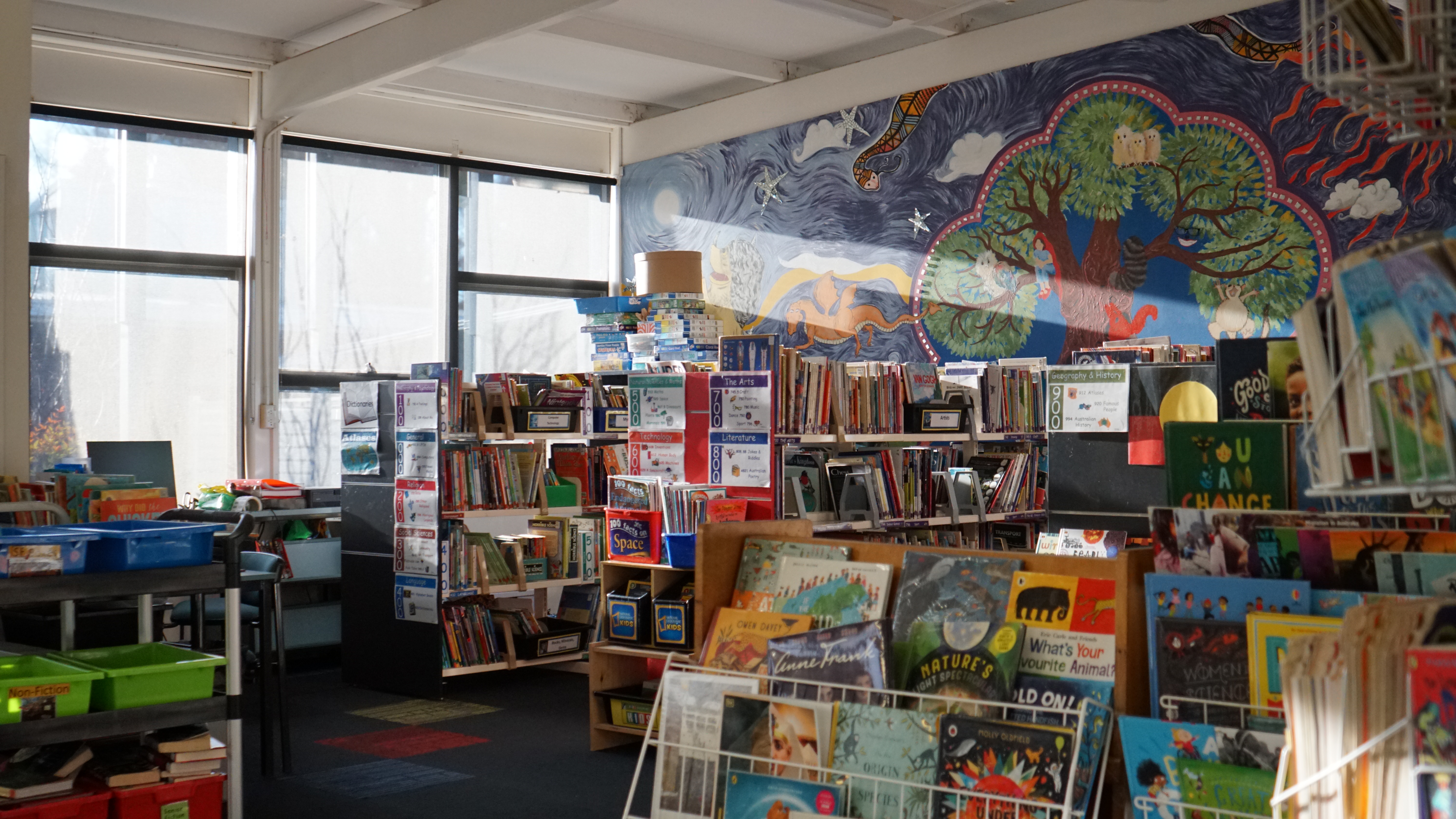 A corner of the library with shelves of books and a tree mural painted on the wall.
